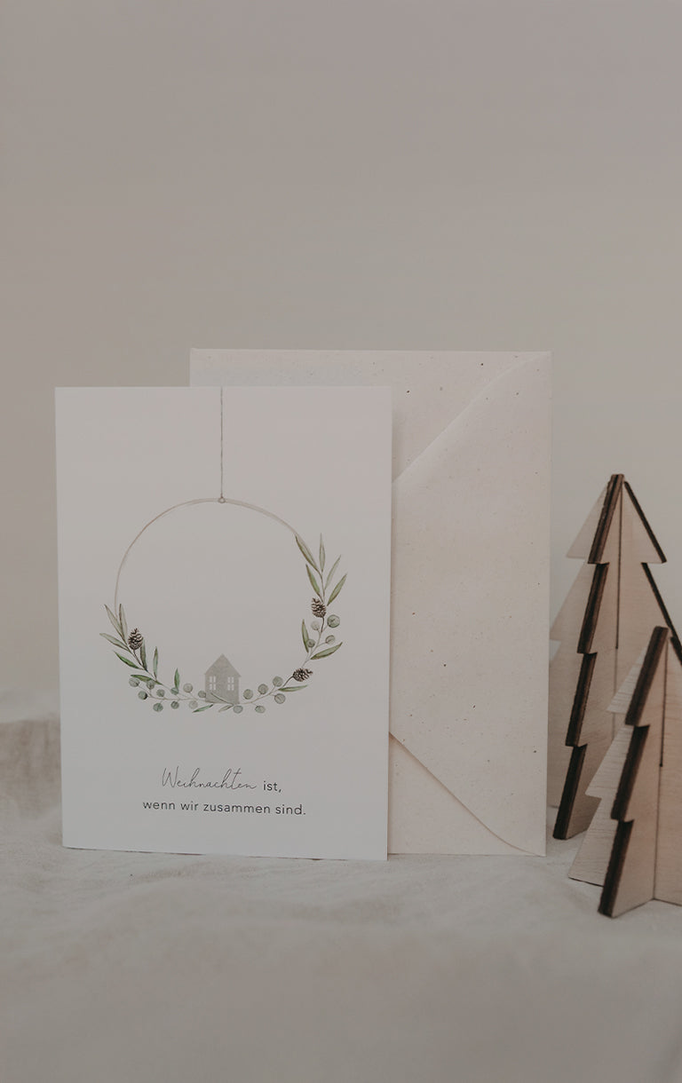 Weiße Klappkarte mit Umschlag. Auf der Karte ein Kranz aus Zweigen und Zapfen mit einem kleinen Haus in der Mitte. Aufschrift: ‚Weihnachten ist, wenn wir zusammen sind.‘ Daneben zwei kleine Holzbäumchen. Darüber der Text: ‚Klappkarten für jeden Anlass‘. Auf der Schaltfläche darunter steht: ‚Klappkarten‘.