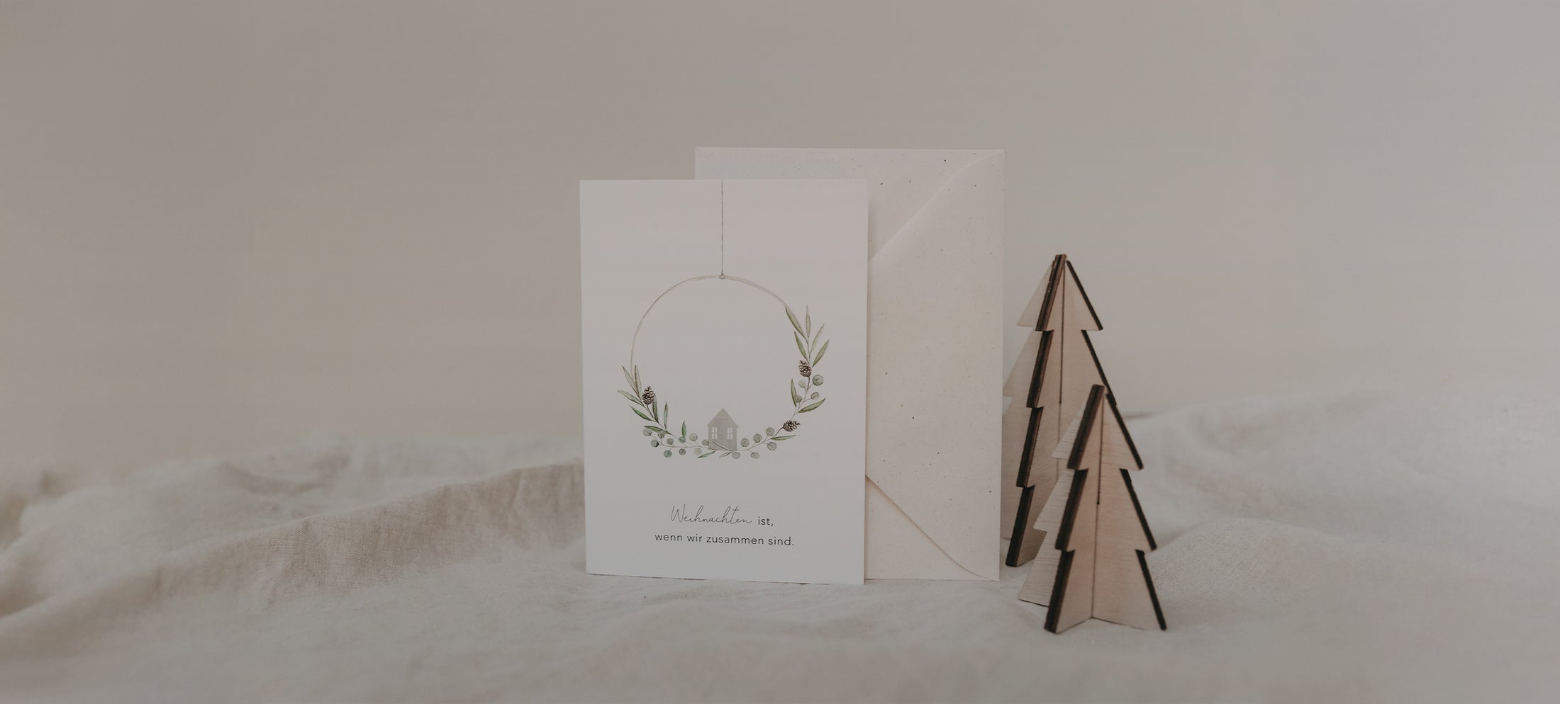 Weiße Klappkarte mit Umschlag. Auf der Karte ein Kranz aus Zweigen und Zapfen mit einem kleinen Haus in der Mitte. Aufschrift: ‚Weihnachten ist, wenn wir zusammen sind.‘ Daneben zwei kleine Holzbäumchen. Darüber der Text: ‚Klappkarten für jeden Anlass‘. Auf der Schaltfläche darunter steht: ‚Klappkarten‘.