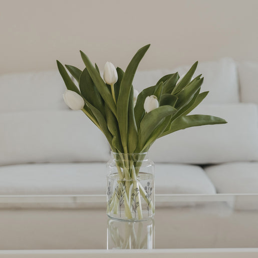 Glasvase mit Blumenwiesen-Motiv und weißen Tulpen auf einem Tisch in heller Wohnumgebung
